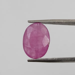 May include: A faceted, oval ruby gemstone, held by tweezers. The ruby is a vibrant pinkish-red colour, with multiple facets catching the light. The gemstone is set against a plain, light background.