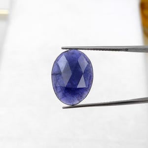 Peut inclure: Une pierre précieuse bleue foncée, taillée en forme ovale, tenue par une pince. La gemme a une surface polie avec de multiples facettes qui reflètent la lumière, créant un effet scintillant. La pierre est d'un bleu riche et saturé.
