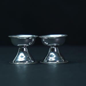 Könnte beinhalten: Zwei silberne Eierbecher mit klassischem Design. Jeder Becher hat eine abgerundete Schale und einen ausgestellten Fuß. Die Becher bestehen aus glänzendem, reflektierendem Metall und stehen vor dunklem Hintergrund.