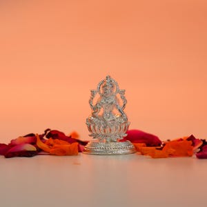 May include: A silver statue of a deity, seated on a lotus flower base, is surrounded by scattered rose petals in shades of red and orange. The background is a gradient of peach and orange.