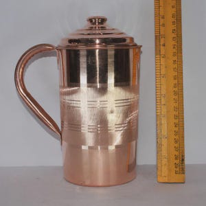 May include: A copper pitcher with a lid and a curved handle. The pitcher has horizontal etched lines and measures approximately 10 inches tall. The pitcher is made of shiny copper and is suitable for serving beverages.