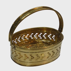 May include: An oval-shaped brass basket featuring a polished metallic finish and a singular arched handle. The sides of the basket are constructed with a decorative pierced cutout pattern, while the base remains solid and smooth.
