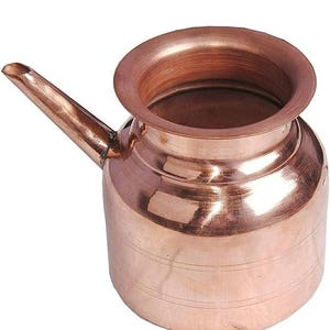 May include: A copper-coloured small pitcher with a spout and a wide, rounded opening. The pitcher has a cylindrical body with horizontal lines and a handle-like spout extending from the side. The metal has a polished, reflective surface.
