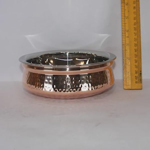 Puede incluir: Un cuenco de color cobre con una textura martillada en el exterior y un borde plateado brillante. El cuenco es redondo y tiene una abertura ancha. Una regla de madera está al lado del cuenco.