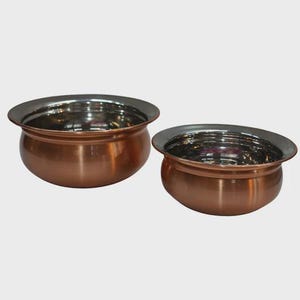 May include: Two round bowls with a polished copper exterior and a smooth, reflective stainless steel interior. The bowls feature a wide, flared rim and a rounded base, showcasing a metallic finish that contrasts the warm copper with silver.