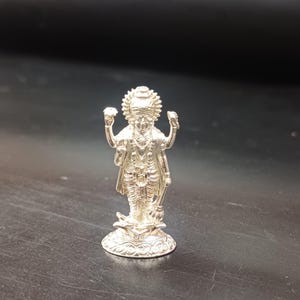May include: A small, detailed silver statue of a deity with multiple arms, wearing ornate clothing and a crown. The statue is standing on a decorative base, with intricate carvings. The background is a dark, neutral color.