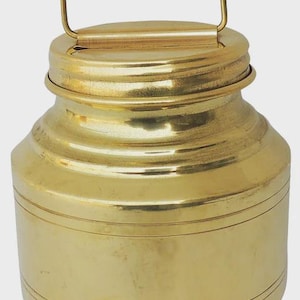 Puede incluir: Un recipiente de metal dorado con asa. El recipiente tiene un cuerpo cilíndrico con bandas horizontales y una tapa. El asa tiene un diseño simple y arqueado. La forma general recuerda a un antiguo lechero.