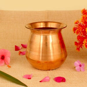 May include: A copper-colored, round pot with a wide rim. The pot is sitting on a tan surface, surrounded by pink flower petals and a few orange flowers. The pot has a shiny, reflective surface.