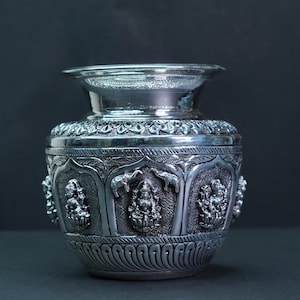 May include: A silver-colored, ornate vase with detailed carvings. The vase features figures and patterns, with a wide base and a flared rim. The design includes figures of deities and elephants. The vase is likely made of metal.