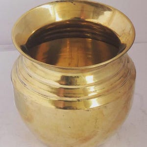 May include: A shiny, golden brass pot with a wide, flared rim and a rounded body. The pot has horizontal ridges around the middle and a smooth, reflective surface. The pot is likely used for cooking or storage.