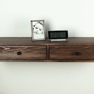 May include: A dark brown, floating wall-mounted shelf with two drawers and black knobs. A framed black and white photo and a digital clock are on top. The shelf is made of wood and has a natural grain pattern.