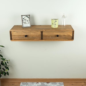 Op de afbeelding: Een zwevend houten plankje met twee lades, elk met een zwarte knop. Een ingelijste foto, een versierd glas en een glazen engelenbeeldje staan erop. Het plankje is aan een witte muur bevestigd.