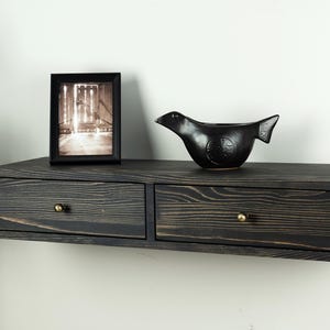 Op de afbeelding: Een aan de muur gemonteerde zwevende plank van donker gebeitst hout met twee lades, elk voorzien van een kleine koperen knop. Op de plank staat een zwarte rechthoekige fotolijst met een zwart-witfoto en een zwart keramieken vogelbeeldje.