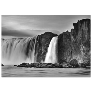 Könnte beinhalten: Schwarz-Weiß-Fotografie eines Wasserfalls, der über felsige Klippen stürzt. Das schnell fließende Wasser erzeugt einen Unschärfeeffekt, der mit den scharfen Details der Felsen und des bewölkten Himmels kontrastiert.