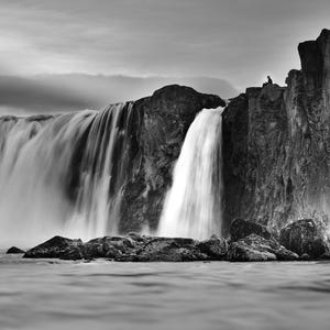 Può includere: Fotografia in bianco e nero di una cascata che precipita su una scogliera rocciosa in uno specchio d'acqua. L'acqua appare liscia, in contrasto con le rocce strutturate e la cascata che scorre. Una persona è in silhouette sulla scogliera.