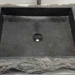 Può includere: Un lavandino rettangolare in pietra grigio scuro con un esterno ruvido e strutturato. Il bacino interno è liscio e scuro, con uno scarico centrale. Una piccola pianta in vaso è visibile sullo sfondo.