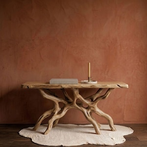 Puede incluir: Una consola de madera natural con una base única en forma de ramas. La superficie de la mesa sostiene un libro, una vela y una pequeña bandeja. La mesa está sobre una alfombra crema contra una pared marrón rojiza.
