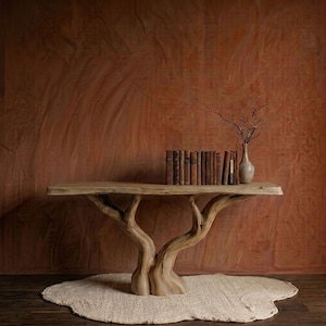 May include: A wooden console table with a tree trunk base. The table holds a collection of vintage books and a small vase with branches. A beige rug sits beneath the table, against a textured, reddish-brown wall.