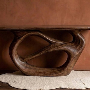Pode incluir: Uma consola de madeira artesanal castanha escura com um design orgânico único. A mesa apresenta uma base escultural com formas abertas e fluidas. Descansa sobre um tapete de cor neutra contra uma parede quente castanho-avermelhada.