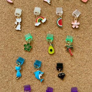 Peut inclure: Une collection de boucles d'oreilles à breloques colorées avec divers motifs, dont un piment rose, un cœur, une licorne, un lion, un chat, un arc-en-ciel, une tranche de pastèque, une fleur, un trèfle à quatre feuilles, un avocat, un palmier, un dauphin, un diamant, une libellule, un éclair et un coquillage.