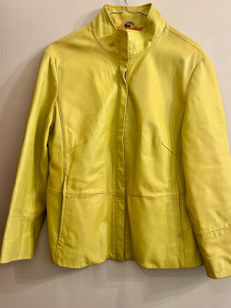 May include: A bright yellow leather jacket with a stand-up collar. The jacket has a tailored fit with seams and panels, and a single-button closure. The sleeves are long, and the jacket appears to be in good condition.