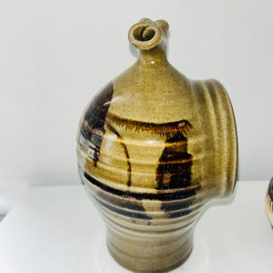 May include: A stoneware jug with a rounded body and a narrow neck featuring a curled spout. The surface has a tan base color with horizontal ridges and dark brown, dripped glaze accents. The vessel has a flat side profile and a circular opening.