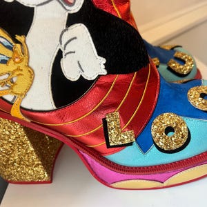 Pode incluir: Uma bota colorida de salto alto com os personagens de desenho animado Piu-Piu e Frajola. A bota tem um salto de glitter dourado, e a palavra "LOVE" é escrita em letras de glitter dourado. A bota é feita de couro vermelho, azul e rosa.