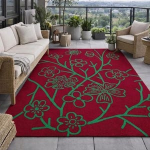 Può includere: Un tappeto rettangolare con uno sfondo rosso intenso e un motivo ripetuto di fiori e rami stilizzati di colore verde brillante. Il tappeto è posizionato su un pavimento in piastrelle grigie davanti a mobili da esterno in vimini.