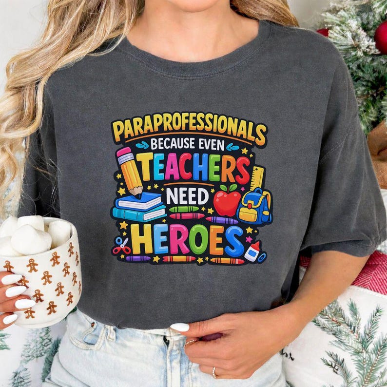 May include: Dark gray t-shirt with a colorful graphic that reads "PARAPROFESSIONALS BECAUSE EVEN TEACHERS NEED HEROES." The design includes a pencil, books, an apple, a backpack, and crayons. The shirt is a comfortable, casual style.