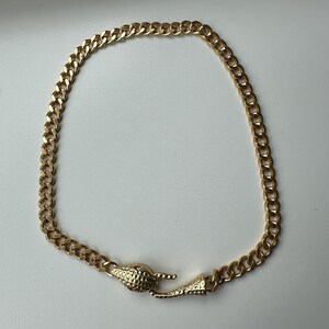 Op de afbeelding: Een goudkleurige ketting bestaande uit een reeks in elkaar grijpende gourmet schakels. De ketting wordt vastgezet met een decoratieve sluiting in de vorm van een gestructureerde slangenkop en staart die in elkaar haken.