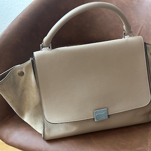May include: A beige leather handbag with a top handle and a flap closure. The bag has suede side panels and a silver-toned metal clasp. The bag is sitting on a brown leather chair.
