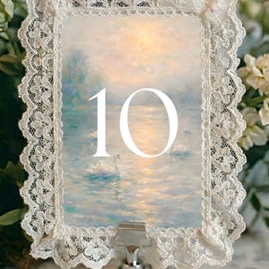 Puede incluir: Una tarjeta decorativa con el número de mesa que presenta una pintura de un lago con cisnes, enmarcada por un delicado borde de encaje blanco. El número "10" se muestra en blanco. La tarjeta está sujeta por un clip de metal plateado.