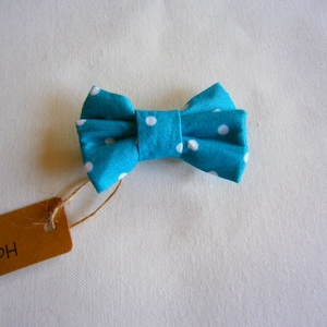 May include: A bright turquoise bow tie featuring a repeating pattern of small white polka dots. A brown rectangular paper tag is attached to the bow tie with a thin piece of twine, with the word Handmade printed on the tag in black ink.