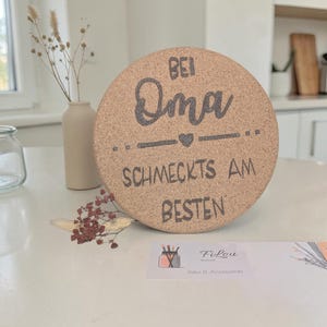 Könnte beinhalten: Ein runder Untersetzer aus Kork mit dem Text "Bei Oma schmeckt's am besten" in Dunkelgrau. Der Untersetzer steht auf einer weißen Oberfläche, im Hintergrund eine kleine Vase und Trockenblumen. Ein dekoratives Küchenaccessoire.
