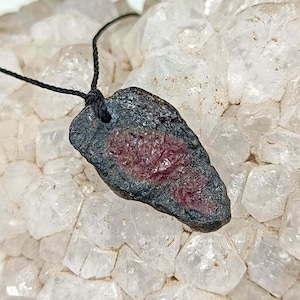 May include: A pendant featuring a raw, dark, textured stone with a deep red centre, suspended from a thin black cord. The stone has an irregular, elongated shape with a visible hole at the top for the cord, showcasing a contrast between the dark exterior and the reddish-maroon interior mineral deposit.