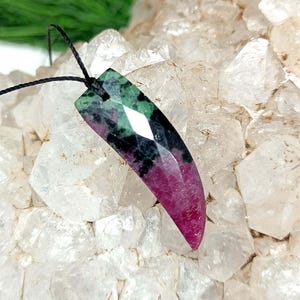 Pode incluir: Um pingente facetado em forma de chifre feito de zoisite com rubi, apresentando uma mistura vibrante de magenta profundo, verde floresta e inclusões minerais pretas. O pingente está suspenso por um cordão preto fino e tem superfície polida.