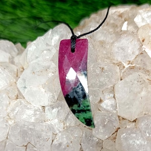 Può includere: Un ciondolo curvo e sfaccettato caratterizzato da una miscela di colori magenta intenso, nero e verde bosco, sospeso a un sottile cordino nero. Il ciondolo presenta una superficie geometrica lucida con bordi affilati e una forma affusolata.