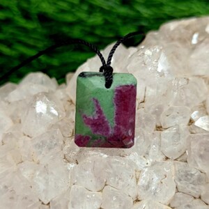 Può includere: Un ciondolo rettangolare con superficie sfaccettata caratterizzato da un mix di motivi minerali color magenta intenso e verde chiaro. Il ciondolo è appeso a un sottile cordino nero e presenta una finitura lucida con distinte variazioni di colore.