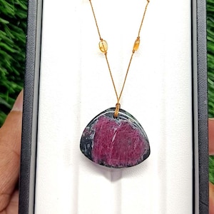 Può includere: Una collana con un ciondolo triangolare dal centro magenta intenso e bordi verde scuro, sospesa a una sottile catenina dorata impreziosita da piccole perline ovali color ambra, presentata in una scatola espositiva rettangolare nera.