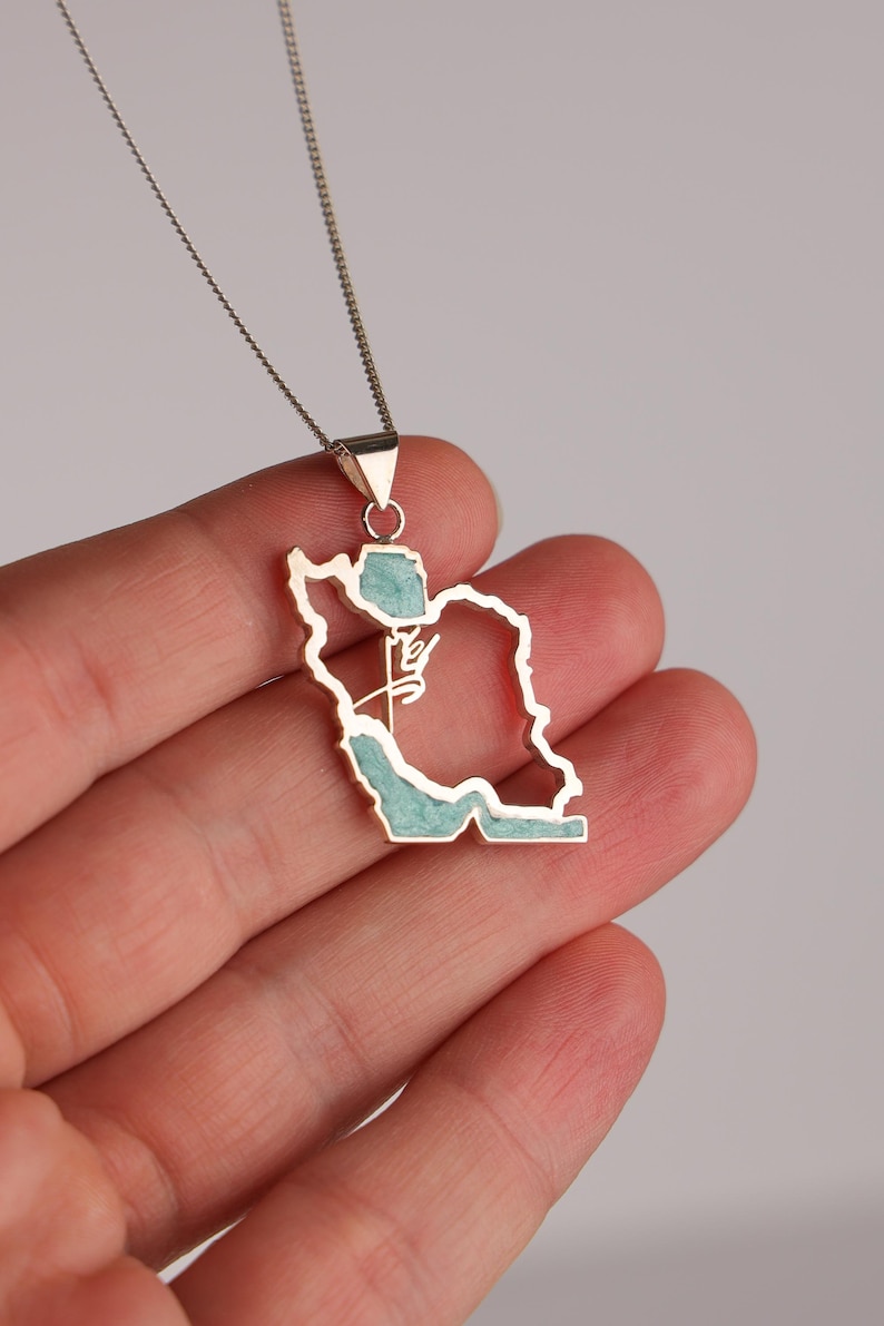 May include: Silver-toned pendant necklace featuring the outline of a country, with a turquoise-colored inlay. The pendant hangs from a delicate chain. The necklace is held in a hand.