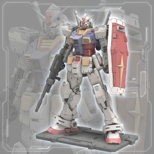 以下が含まれることがあります： ガンダムロボットの詳細なモデル。主に白で、赤、青、黄色のアクセントがあります。黒いライフルと金色の星が付いた赤いシールドを持っています。ロボットは灰色のプラットフォームに立っており、背景にはロボットのぼやけた輪郭があります。