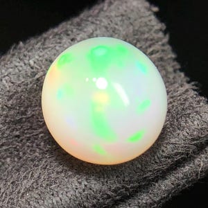 Peut inclure: Une pierre précieuse opale ronde taillée en cabochon, présentant une couleur de base blanc laiteux avec des éclats iridescents vibrants de vert néon et des nuances subtiles de lumière orange et jaune sur sa surface lisse, polie et bombée.