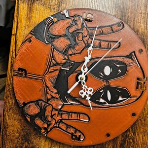 May include: Circular wooden wall clock with an orange and black graphic design. The clock face features a stylized illustration of hands making the peace sign and a character's mask. The clock hands are white.