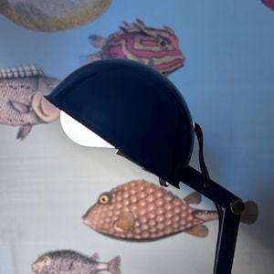 Op de afbeelding: Een marineblauwe bureaulamp met een ronde kap en verstelbare arm. De lamp staat voor een lichtblauwe muur versierd met illustraties van diverse kleurrijke vissen.