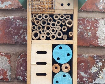 Könnte beinhalten: Insektenhotel aus Holz mit hellblauem Dach und Akzenten, an einer Ziegelmauer befestigt. Das Haus verfügt über verschiedene Fächer, darunter ein Drahtgeflechtbereich, Bambusröhren und gebohrte Holzblöcke.