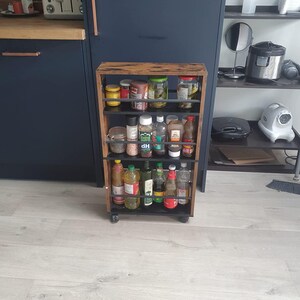 Puede incluir: Un especiero de madera sobre ruedas, lleno de varios frascos y botellas de especias y condimentos. El especiero tiene cuatro estantes y un marco de metal oscuro. La madera tiene un acabado natural. El especiero está en un entorno de cocina, con un armario azul oscuro en el fondo.