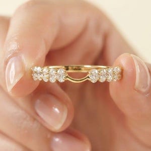 May include: A gold ring guard featuring a row of small, clear, oval-cut stones set along the band, with a distinct curved gap in the center designed to accommodate another ring. The metal band has a polished, reflective yellow gold finish.