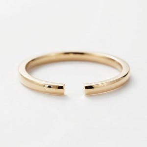 May include: A polished gold-tone open ring featuring a smooth, rounded band with a small gap between the two flat, squared-off ends. The metal has a reflective surface and a consistent, uniform thickness throughout the circular design.