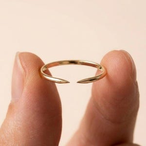 Può includere: Un delicato anello d'oro con un design aperto unico. L'anello presenta una fascia sottile che si divide nella parte superiore, formando due estremità appuntite. L'anello è tenuto tra le dita su uno sfondo morbido e neutro. L'anello è contrassegnato con "14K".