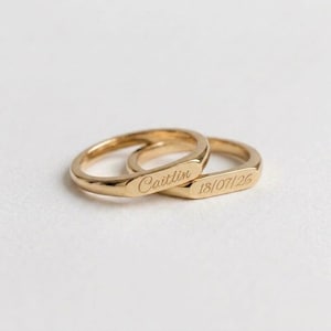 Puede incluir: Dos anillos de tono dorado con un acabado liso y pulido. Un anillo presenta el nombre "Caitlin" grabado en letra cursiva, mientras que el otro anillo muestra la fecha "18/07/26" grabada en una fuente sans-serif clara sobre una superficie plana.